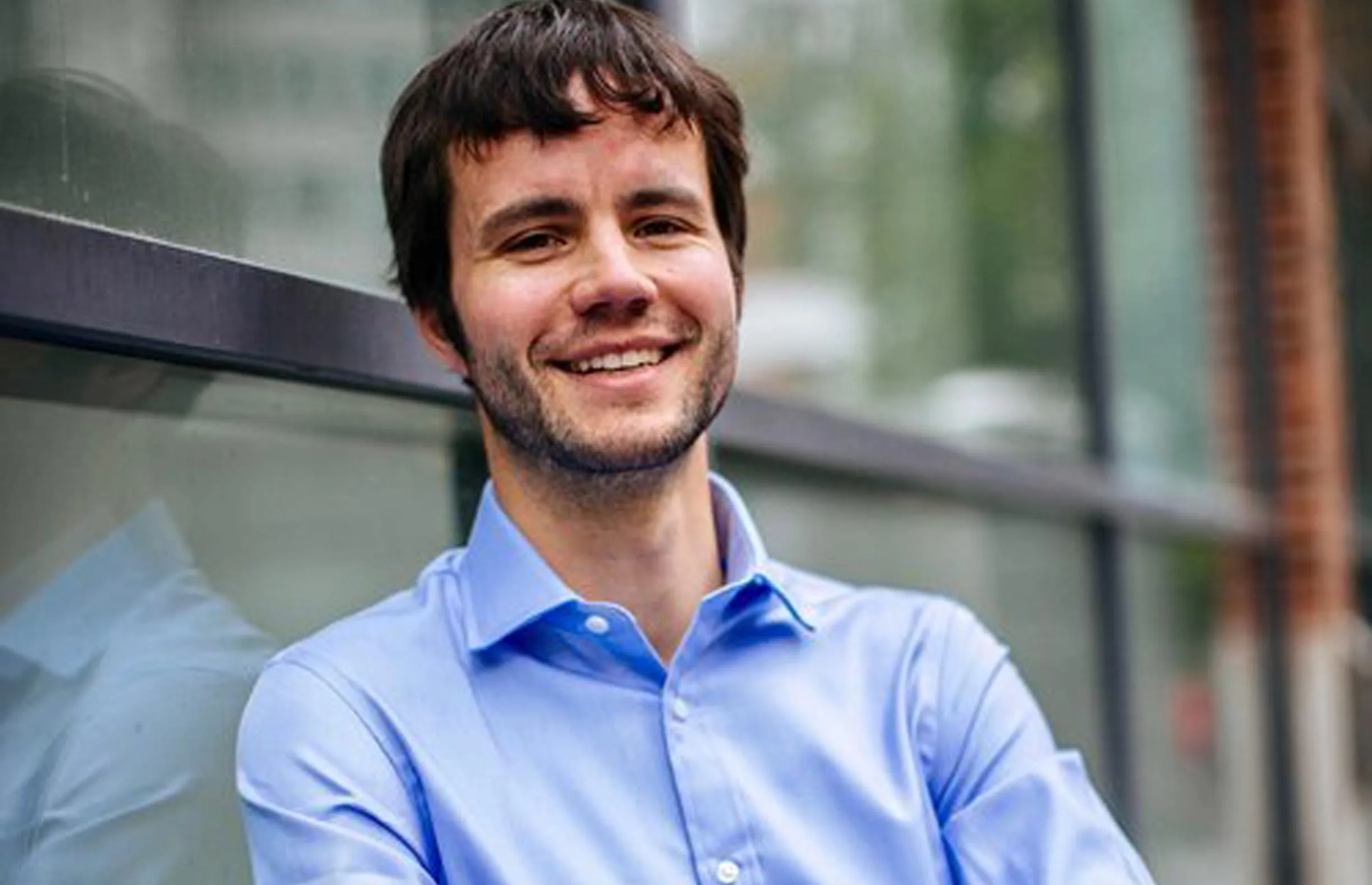 Sebastian Bock leans against a window outside a building at LSE, He is wearing a light blue shirt and smiles at the camera.